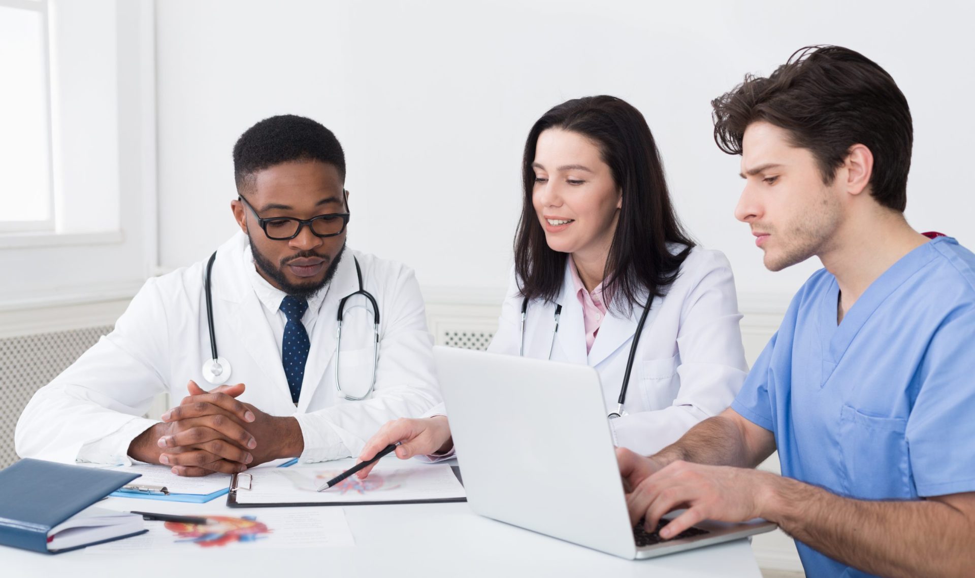 Bilden visar tre personer med medicinrock och medicinsk utrustning, en laptop samt en bok som sitter vid ett bord och samtalar.