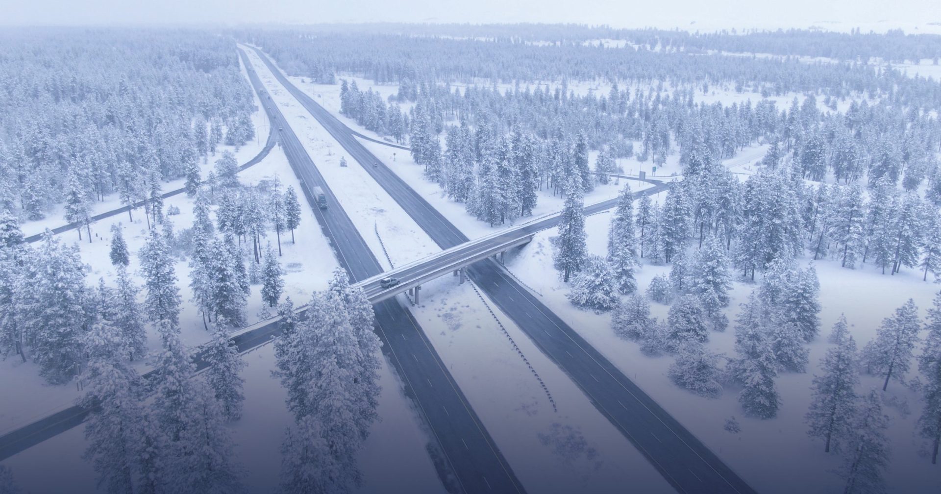 i ett snöigt och dimmigt landskap ser man en bro och vägar ovanifrån. Omgärdat finns granar som är snöklädda.
